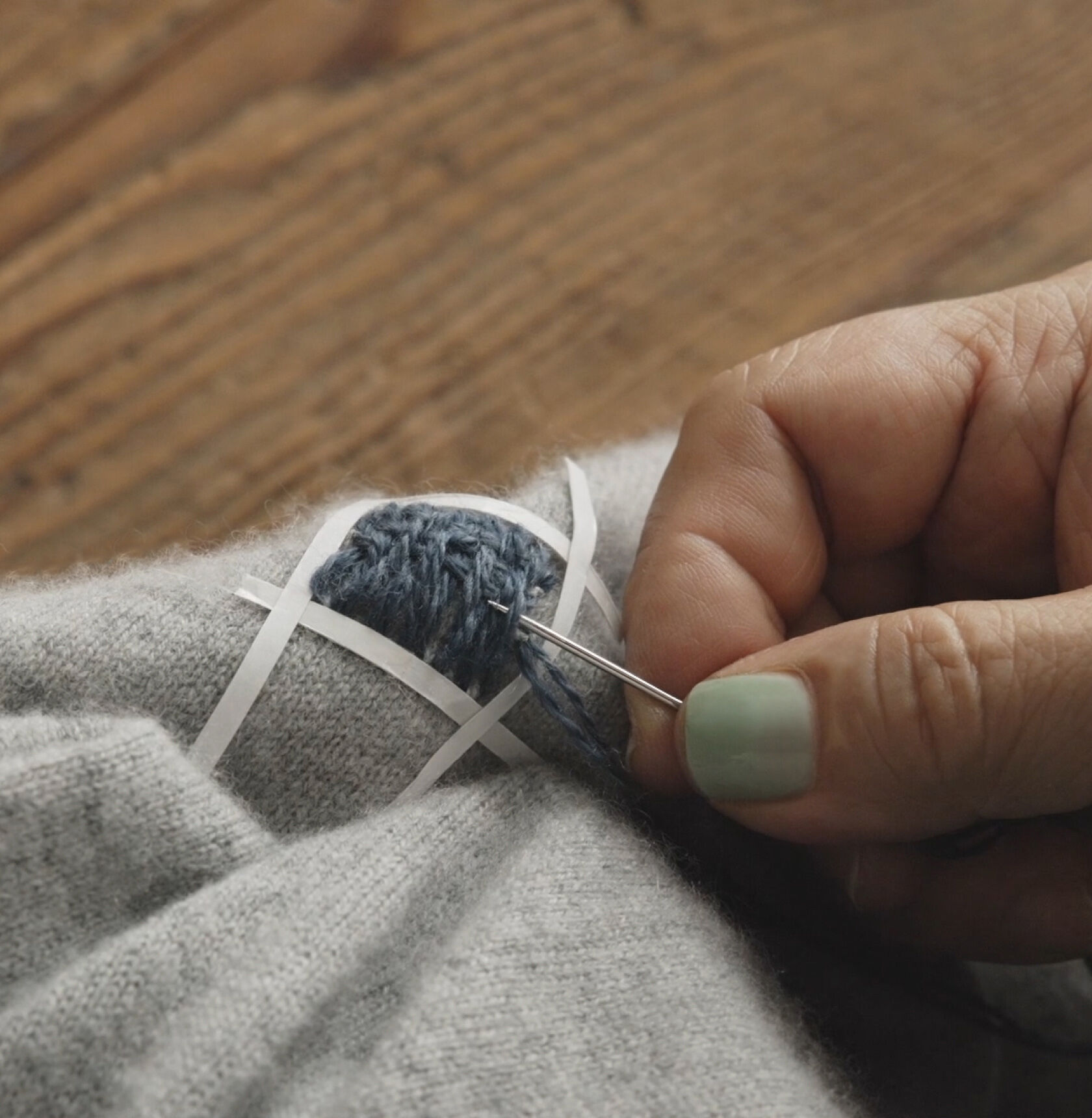 Close-up of hands weaving blue thread horizontally on a mended gray sweater. Close-up of hands weaving blue thread horizontally on a mended gray sweater.